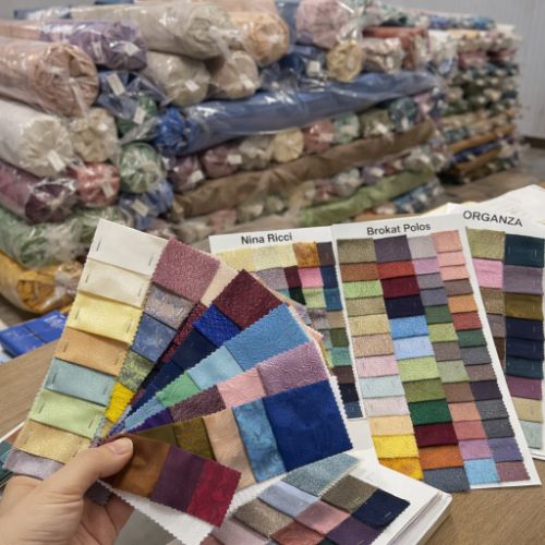 Swatch kain pelbagai jenis di atas meja dengan sampel potongan kain dan timbunan roll kain di belakang untuk carian material pakaian.