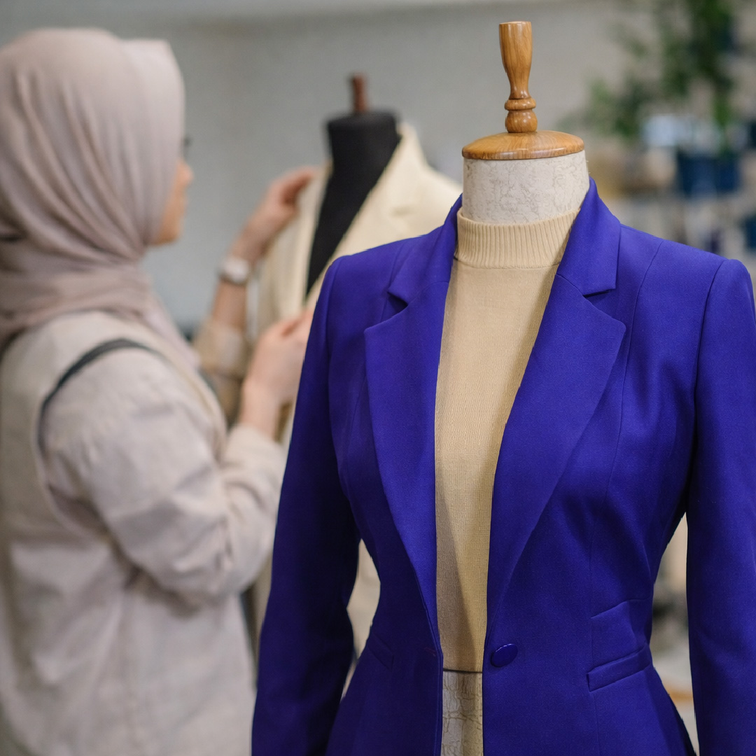 Mannequin memakai blazer wanita siap jahit dengan latar belakang designer sedang fitting sebagai proses penyediaan sampel pakaian